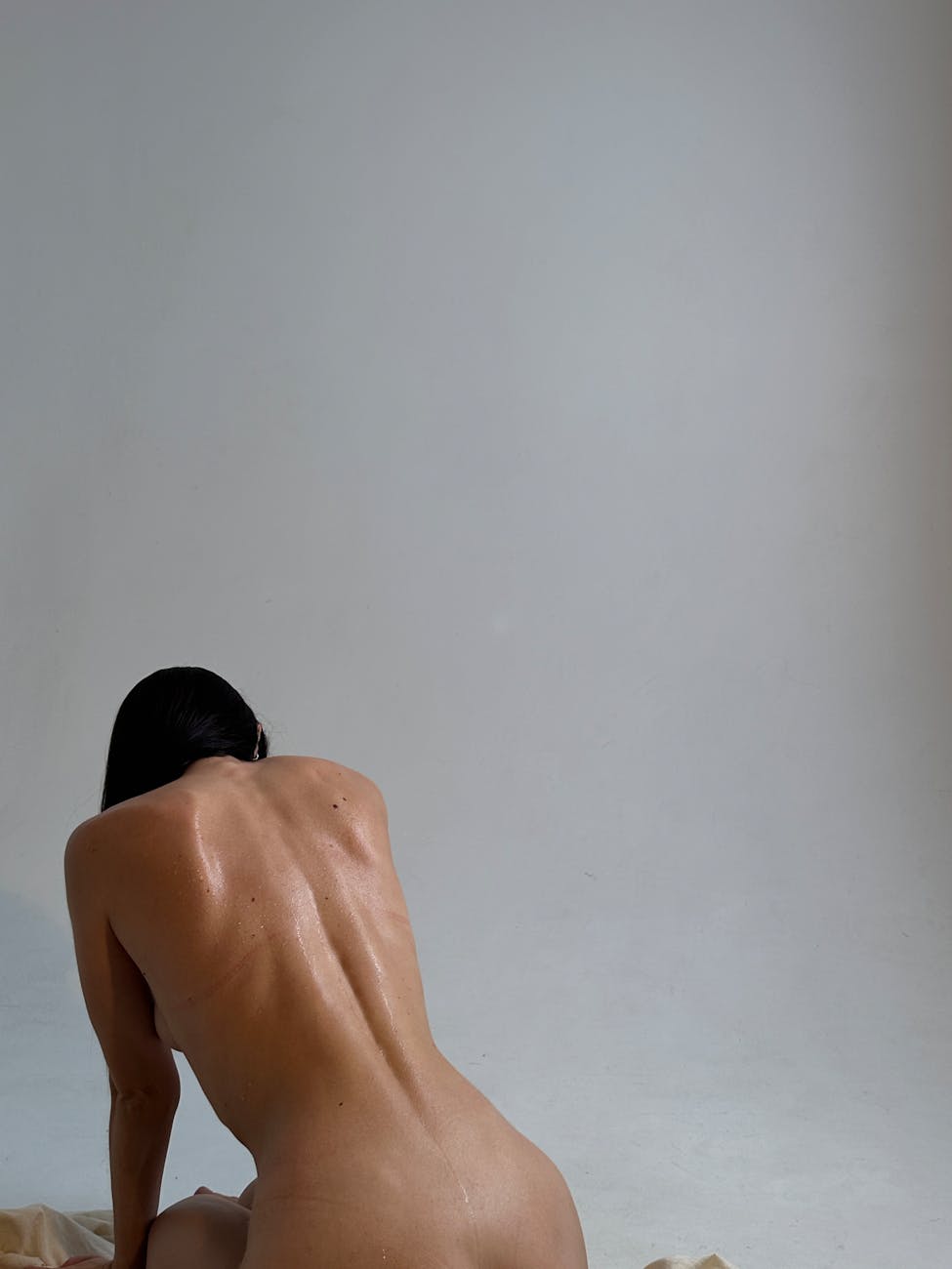 A back view of a woman posing nude in a minimalist studio setting with a white background.